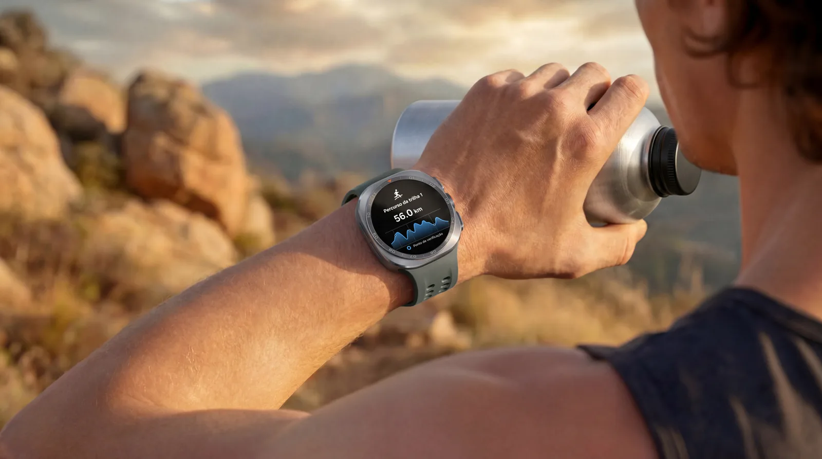Corredor bebe água numa trilha com o Galaxy Watch Ultra2 no pulso; a tela em português mostra o Percurso da trilha 1, com 56 km